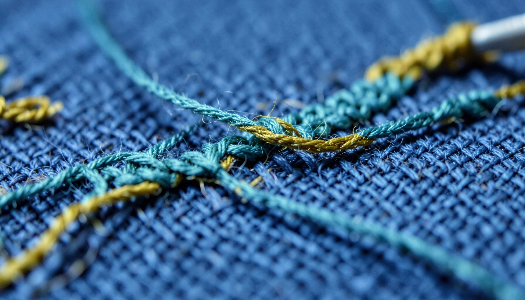 Extreme close-up of needlework thread in muted teal and mustard looping over linen weave